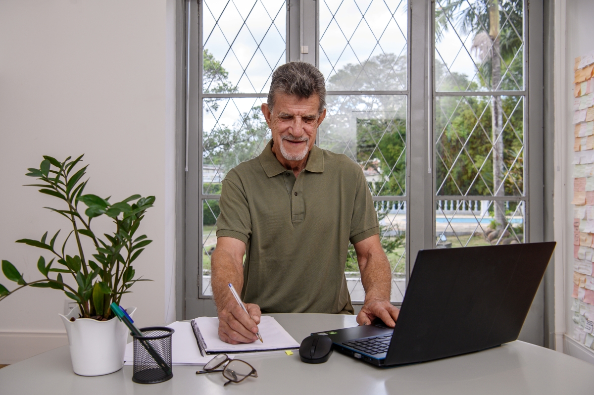 Um homem sorrindo sentada em frente ao notebook enqaunto escreve no caderno.