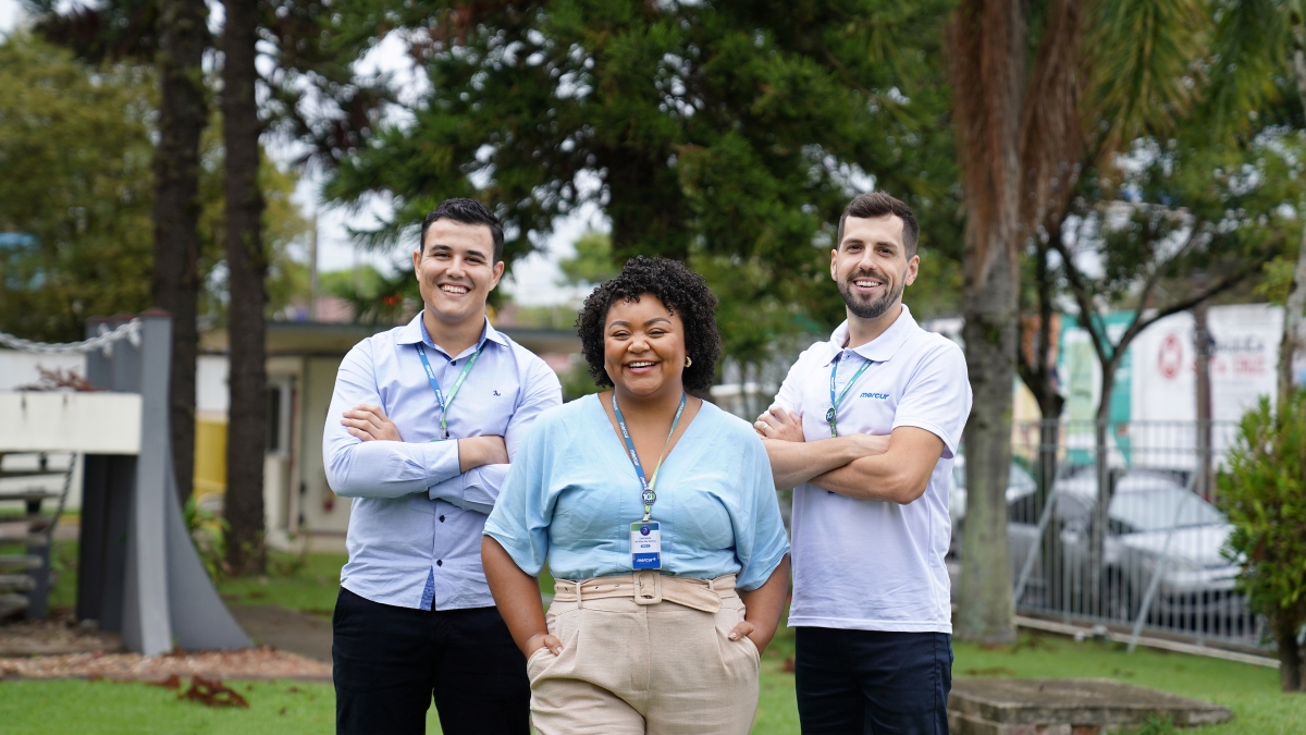 3 pessoas colaboradoras da Mercur, dois homens sorrindo com os braços cruzados e ao centro, uma mulher sorrindo com as mãos no bolso.