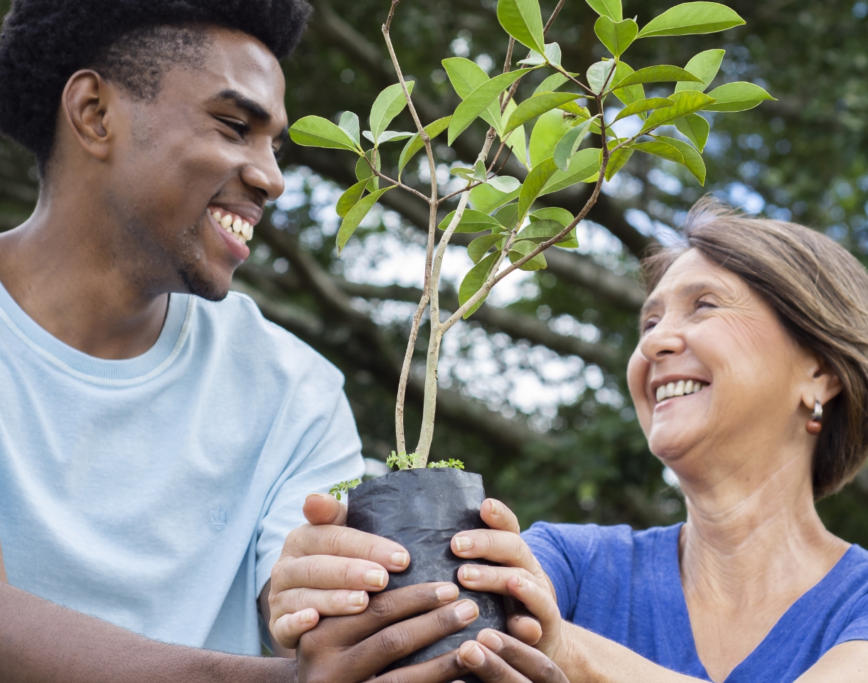 Um homem jovem e uma mulher idosa olhando um para o outro e sorrindo enquanto seguram uma muda de planta.