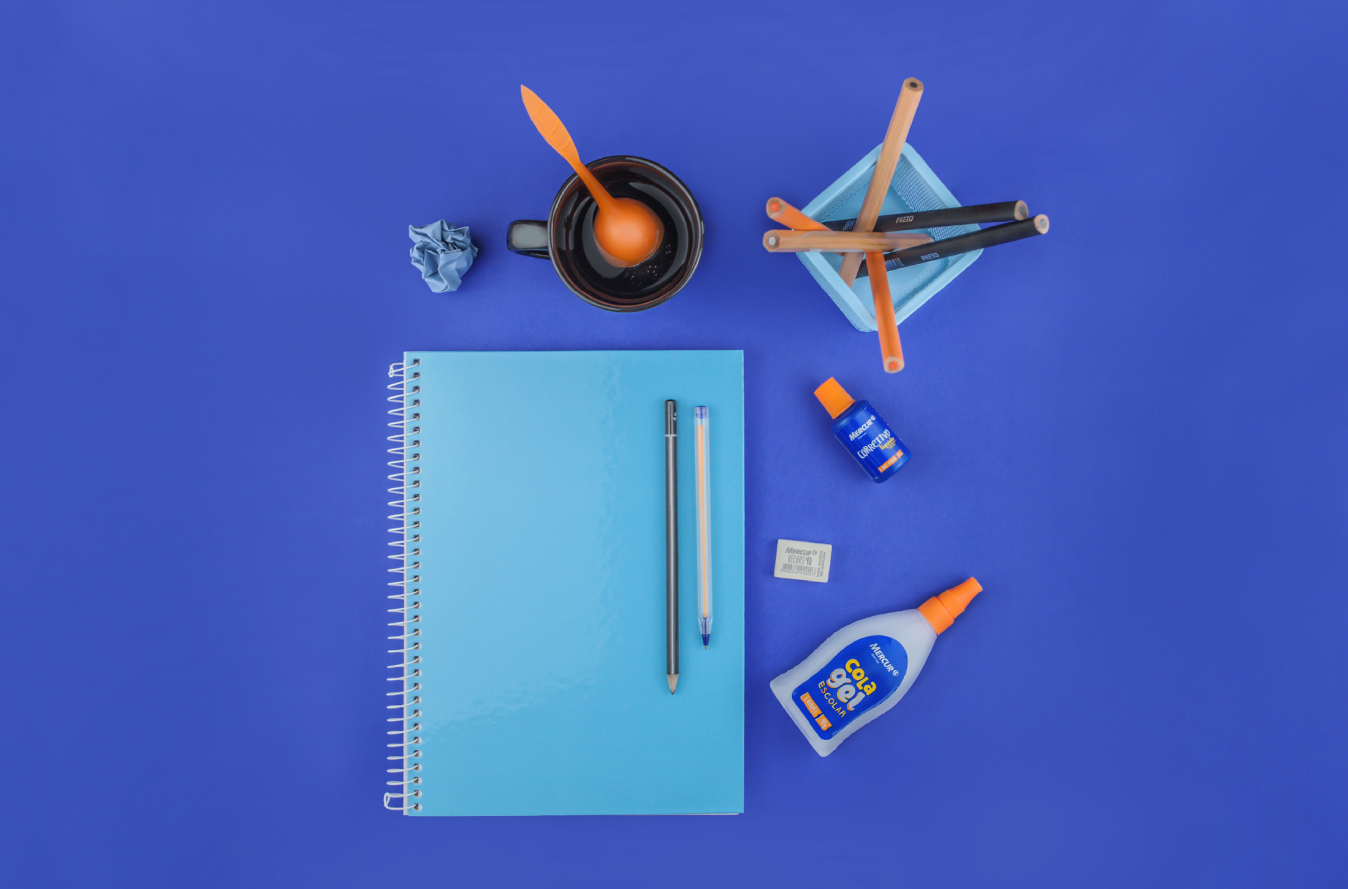 Sobre uma superfície azul, há diversos materiais escolares vistos de cima: um papel azul claro amassado, um porta lápis com seis lápis dentro, um caderno de cor azul clara, com um lápis e uma caneta sobre ele, um corretivo uma borracha e a Cola Gel da Mercur.