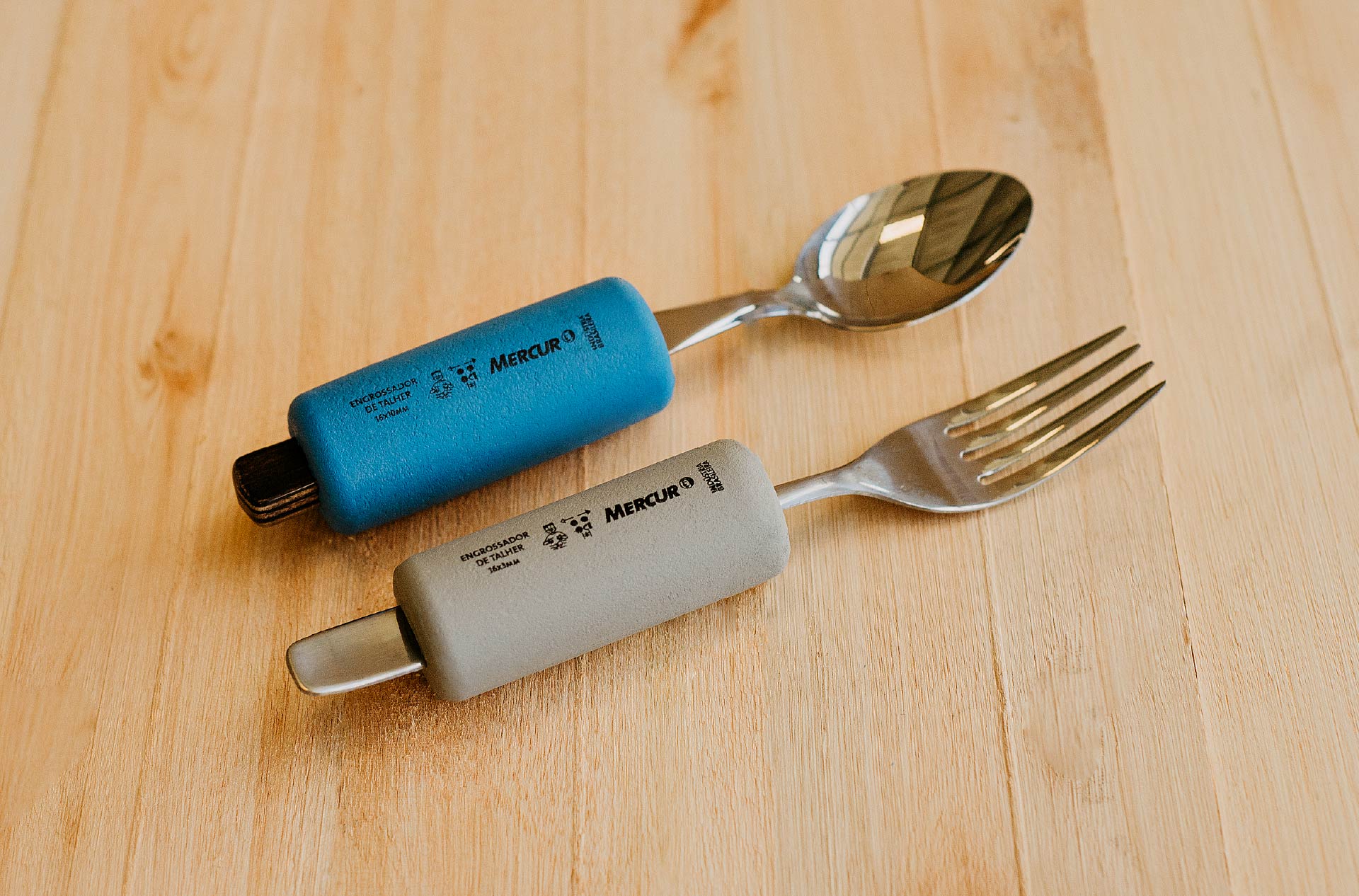 Em uma mesa de madeira, o Engrossador de Talheres azul envolve uma colher com cabo de madeira e o Engrossador de Talheres cinza envolve um garfo com cabo de metal.