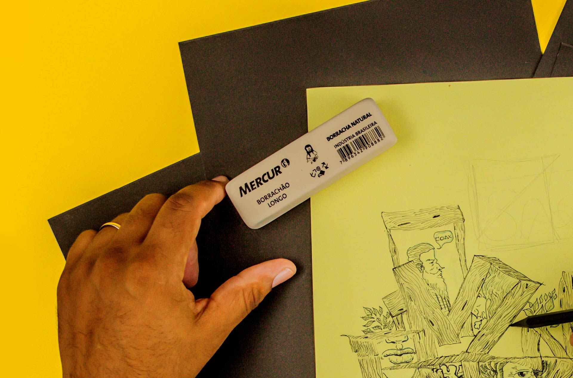 Vemos de cima as mãos de uma pessoa fazendo um desenho abstrato de lápis em uma folha amarela. Acima do desenho, está o Borrachão Longo, uma borracha branca, de forma retangular, de tamanho grande.