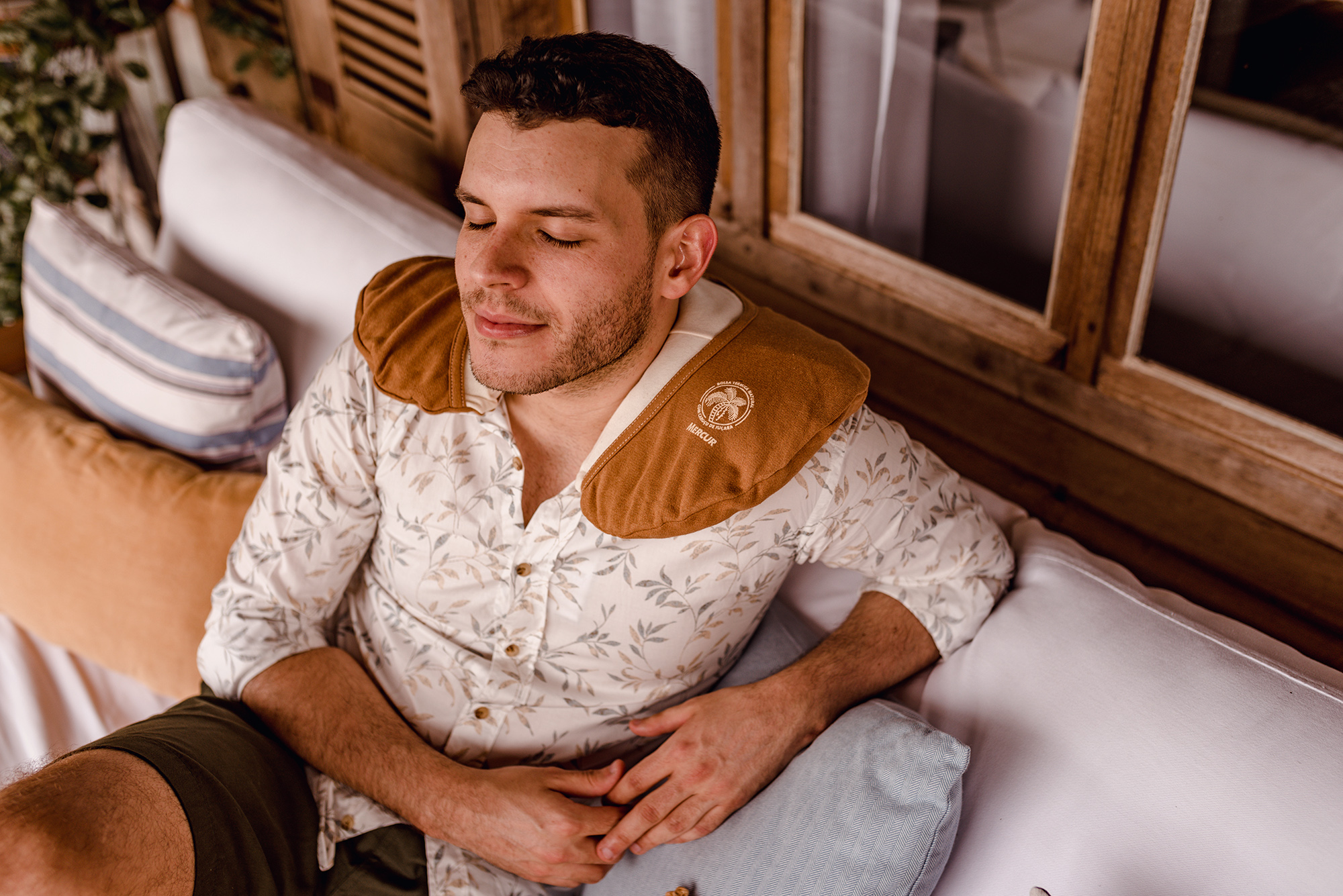 Um homem está sentado em um sofá, com os olhos fechados e expressão relaxada. Nos ombros ele utiliza a Bolsa Térmica Natural Cervical.