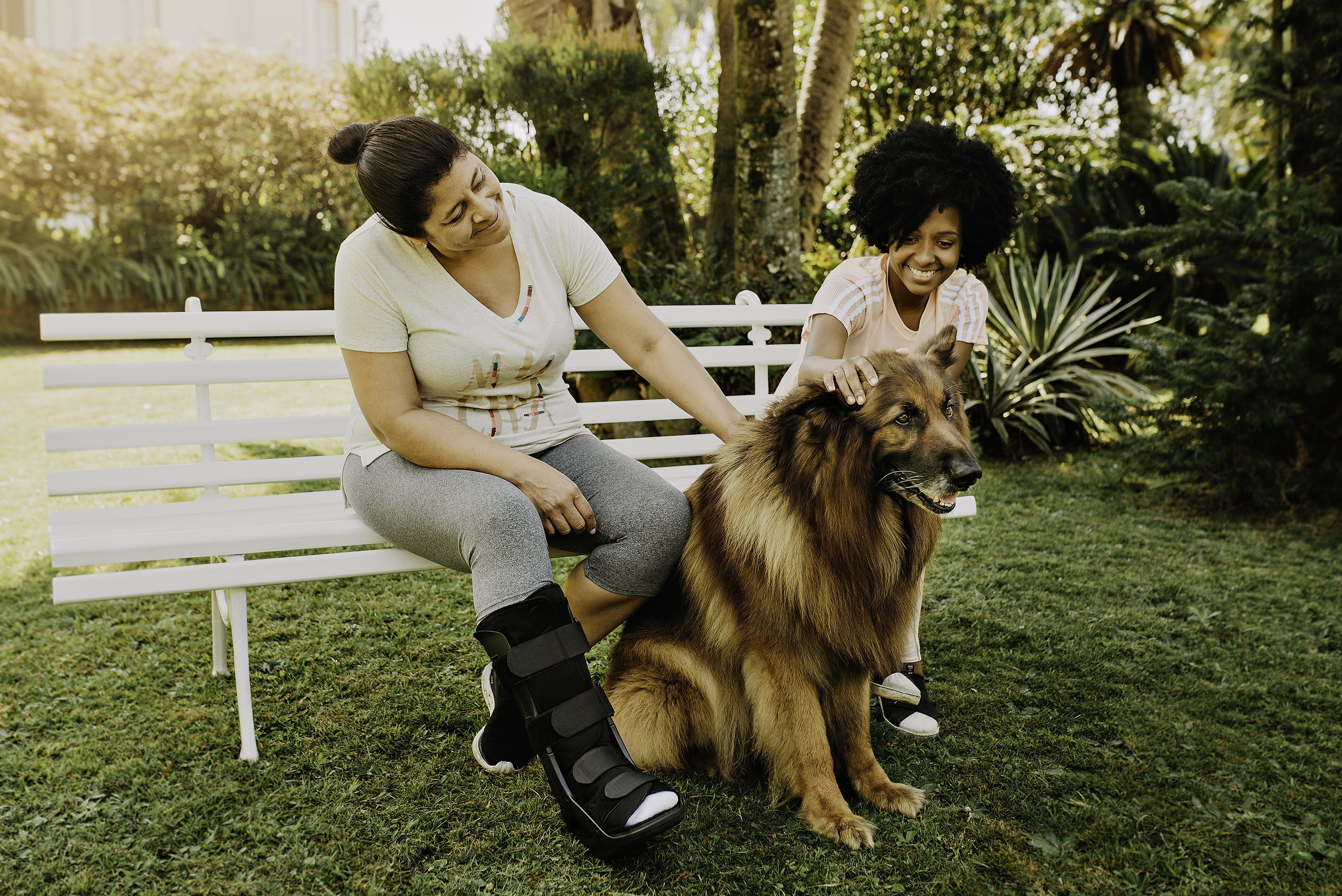Duas mulheres estão sentadas lado a lado em um banco, ao ar livre. Elas estão sorrindo e acariciando um cachorro de grande porte que está sentado em frente a elas.
