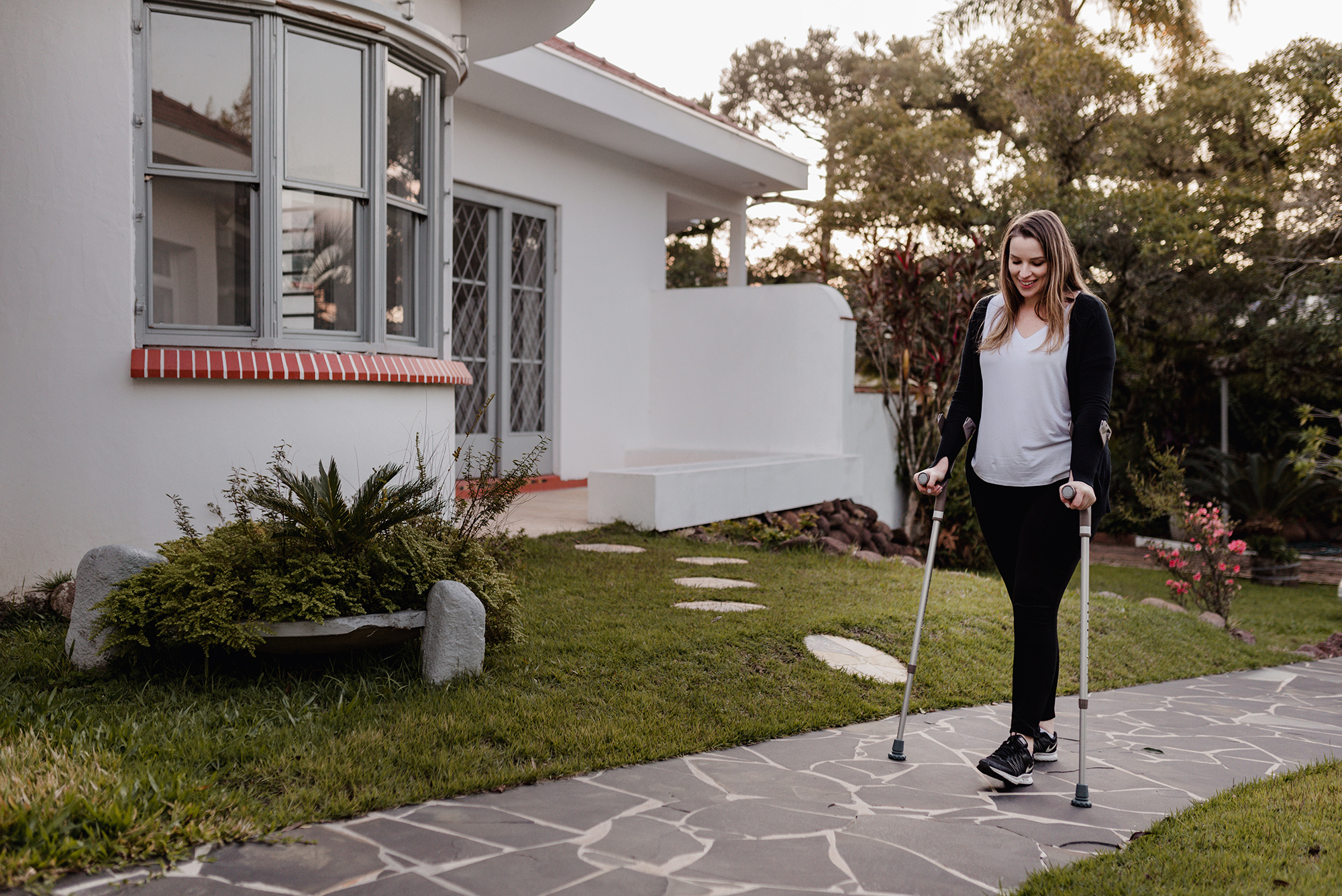 Uma mulher está caminhando com o auxílio de duas Muletas Canadenses Fixas. Ela está em um jardim e sorri, enquanto olha para o lado esquerdo da foto. 