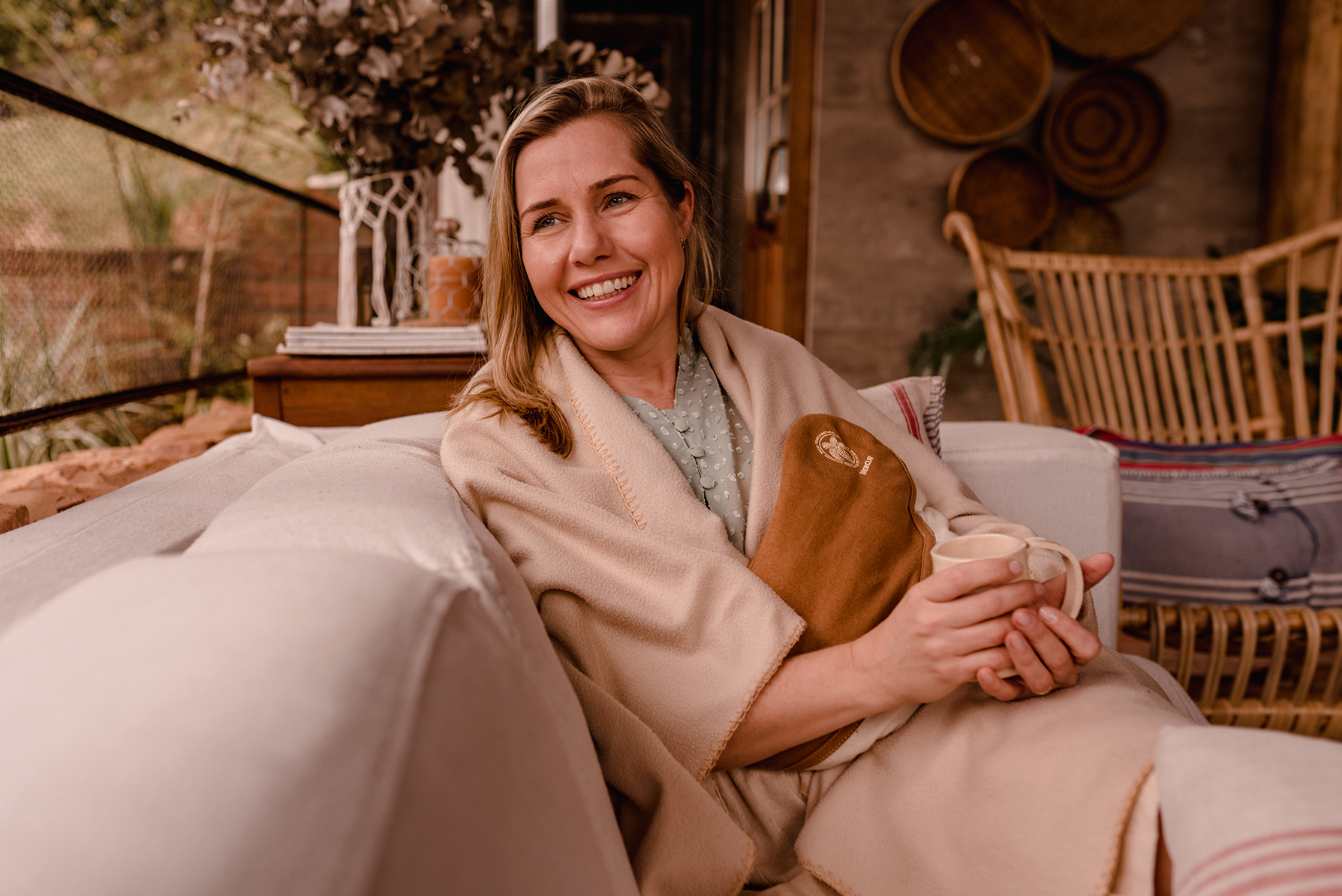 Uma mulher está sentada em um sofá envolta em uma coberta, segurando uma xícara e utilizando uma Bolsa Térmica Natural Grande sobre seu abdômen. Ela sorri, olhando para o lado esquerdo da foto.