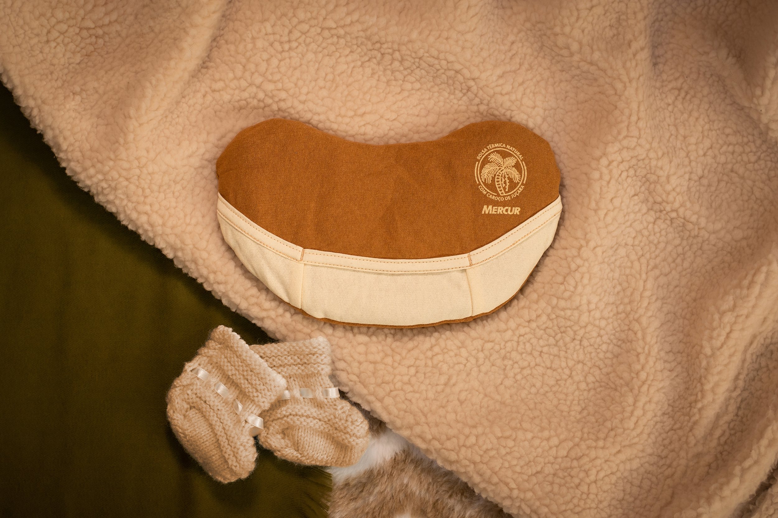 Imagem vista de cima de uma Bolsa Térmica Natural pequena, disposta sobre alguns cobertores. Abaixo, à esquerda da Bolsa, um par de sapatinhos de bebê, em tricô.