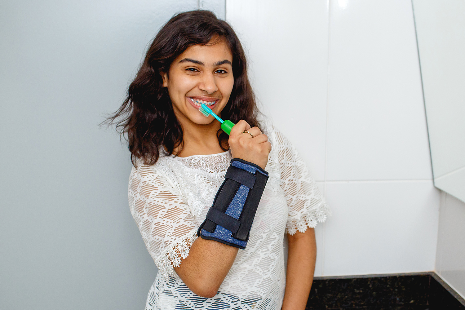 Uma adolescente sorridente segura uma escova de dentes com um engrossador multiuso verde. No seu punho e antebraço, uma pulseira de peso permite que ela segure a escova de dentes com firmeza.