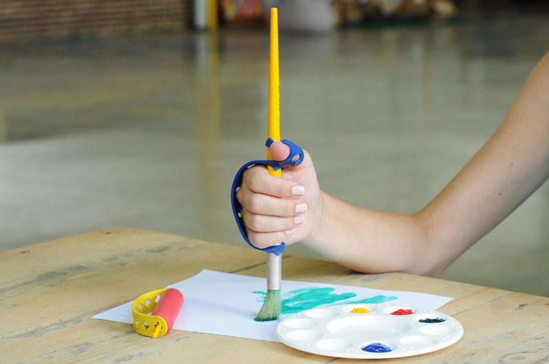 Uma mão feminina segura um pincel amarelo com a ajuda de um fixador em tira na cor azul. Ela está pintando uma folha em branco e ao lado tem um recipiente com tintas nas cores azul, amarelo, vermelho e preto.