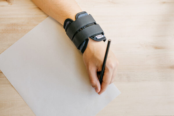 Sobre uma folha branca e segurando um lápis, uma pessoa escreve com a ajuda de uma pulseira de peso da Mercur.