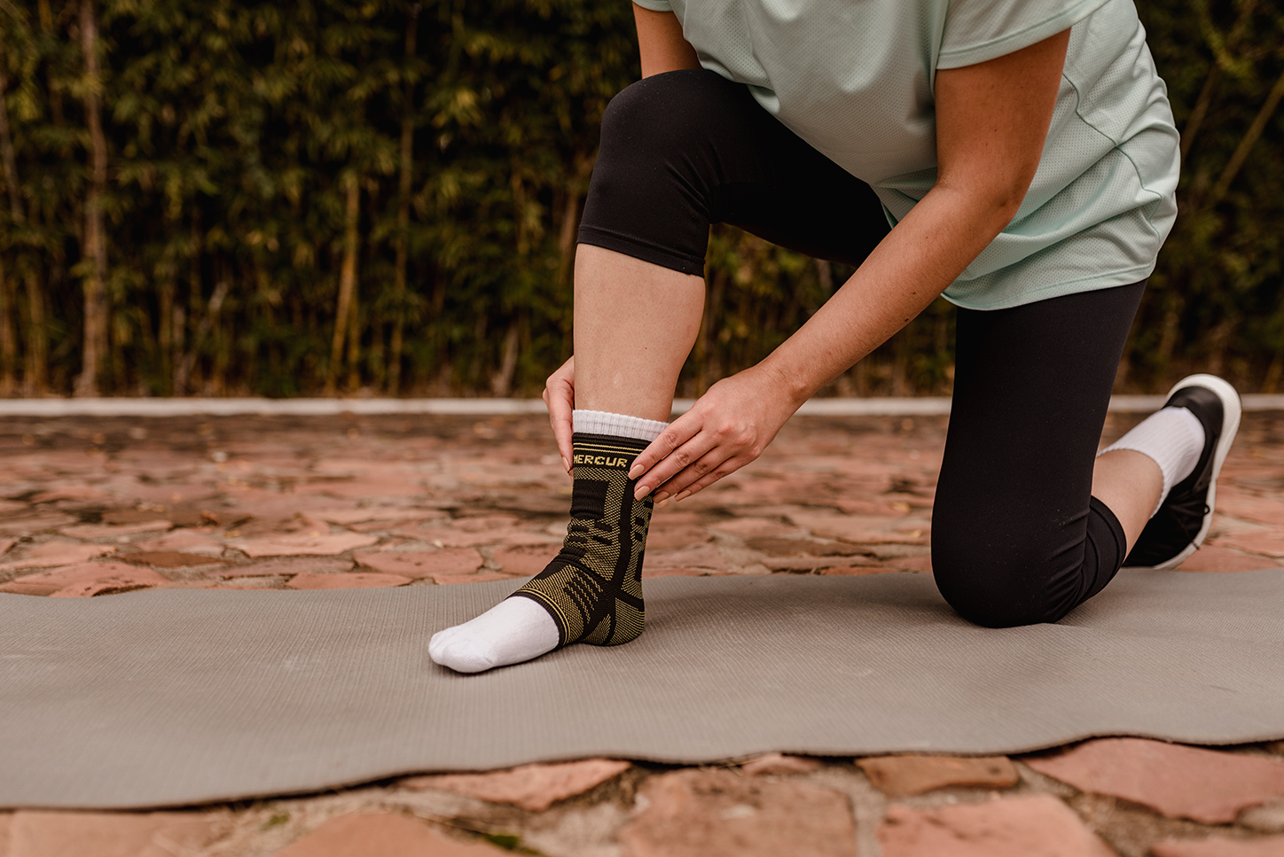 Uma mulher está ajustando a Tornozeleira de Compressão Elástica da Mercur, preta com verde no seu pé direito que está apoiado em um tapete de yoga. Seu joelho esquerdo está no chão para dar apoio. 